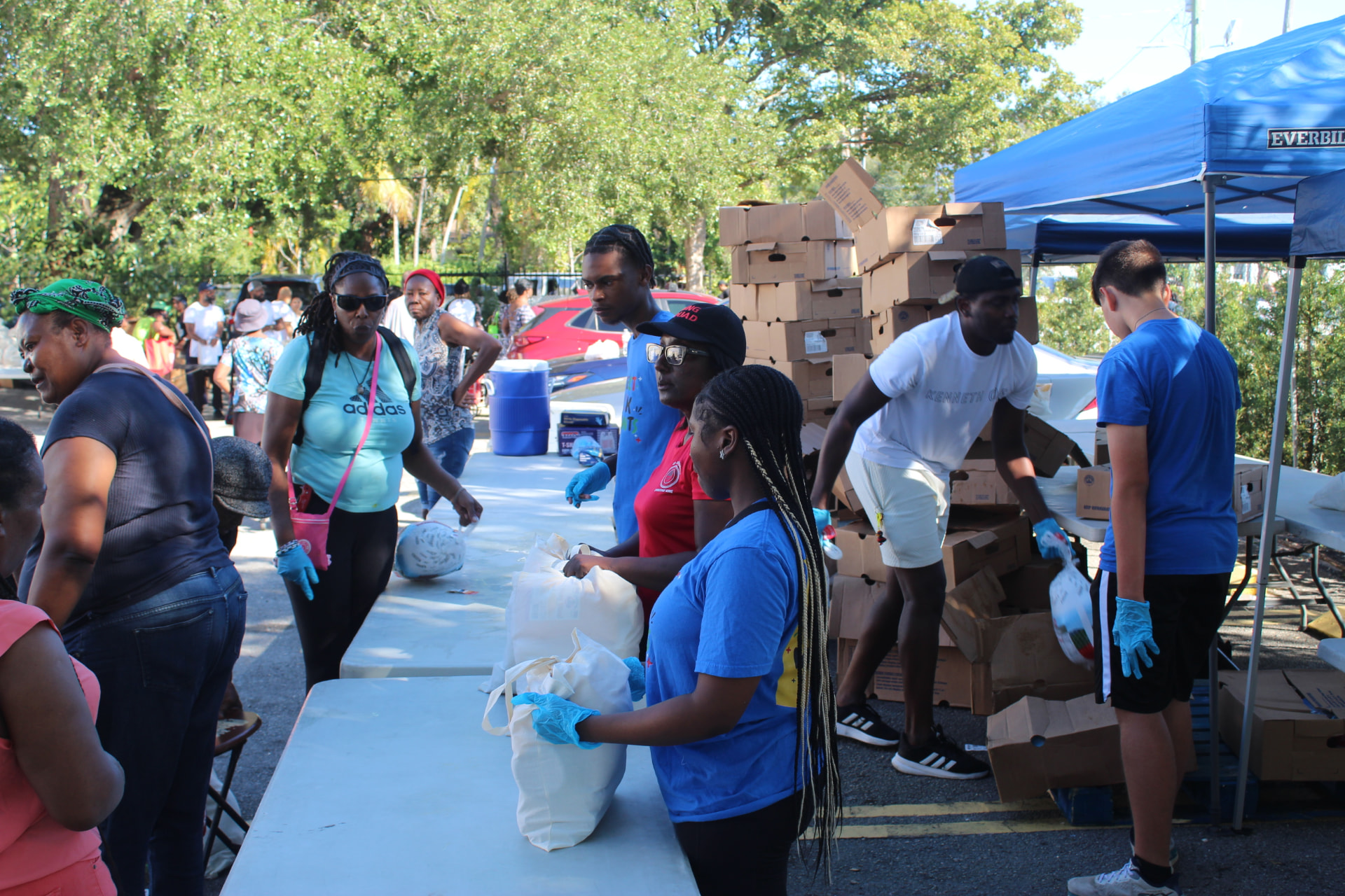 Volunteers in blue gloves packing bags of groceries at a TAPARI food distribution under blue canopies